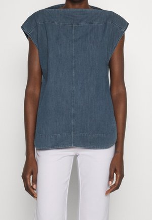 Osoba ubrana w niebieski jeansowy top bez rękawów z wyściełanymi ramionami i białe spodnie, stojąca na jednolitym tle.