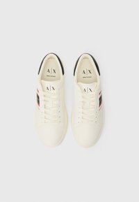 White synthetic lace-up sneakers featuring black heel accents and red and black stripes on the sides. Textured surface and logo on the tongue.