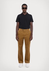 Schwarzes Poloshirt, braune Cordhosen und weiße Sneakers. Runder Kragen, Knopfdetails an der Hose. Model steht vor einem schlichten Hintergrund.