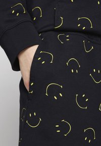 Tissu noir à la texture lisse, orné d'un motif répétitif de visages souriants jaunes. Comprend une poche à la taille.