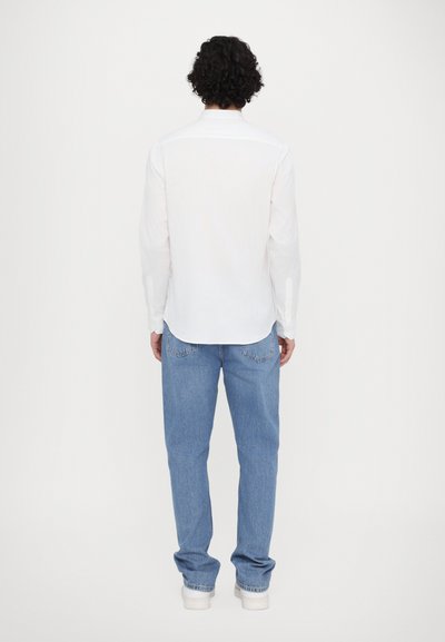 Hvit langermet skjorte med bakstykke og buet kant, kombinert med lyseblå denimjeans med klassisk passform og synlige baklommer.