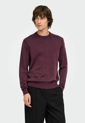 Man with medium-length brown hair wearing a long-sleeve maroon sweater and black pants looking to the side with one hand in his pocket.