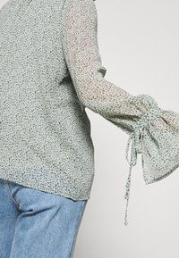 Grön mönstrad blus i genomskinligt tyg, med draperade långärmar och knytband vid ärmsluten. Bärs med ljusblå denim jeans.