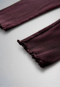 Extremos de las mangas de una prenda de punto morado oscuro con detalle de dobladillo ondulado, extendida sobre una superficie gris clara.