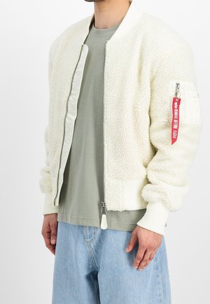Mand iført en cremefarvet sherpa bomberjakke, salviegrøn t-shirt og lyseblå jeans. Rødt mærke hænger fra jakkens ærmelomme.