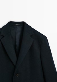 Schwarzer Anzugblazer mit Reverskragen und Einzelknopfverschluss, von der Brust aufwärts vor einem weißen Hintergrund gezeigt.
