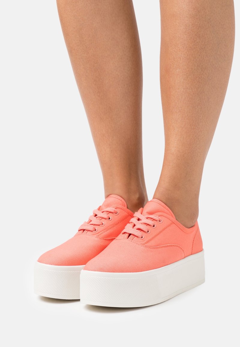 Jambes portant des baskets compensées rose corail avec des semelles blanches épaisses, debout sur un fond clair uni.