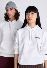 Du modeliai, apsirengę baltais Lacoste džemperiais su gobtuvu, vienas su pilku beanie, pozuoja prieš pilką baltą fono.