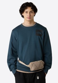 Felpa teal con patch logo e colletto a coste, indossata sopra una camicia beige. Marsupio beige con superficie testurizzata indossato in vita.