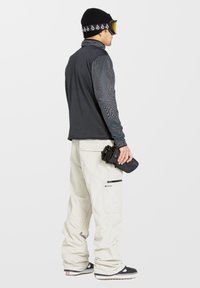 Zwart en grijs patroon op een lange mouwen shirt, witte sneeuwbroek met zakken, en zwarte handschoenen. De persoon draagt een zwarte beanie en goggles.