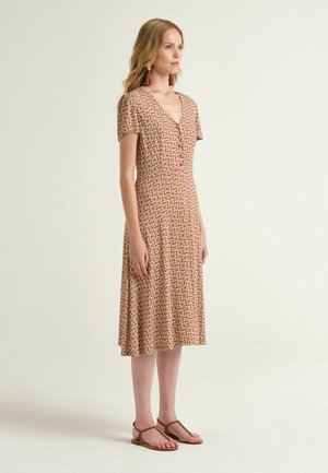 Jeune femme portant une robe midi à manches courtes et motif, avec un décolleté en V, chaussée de sandales plates marron et de boucles d'oreilles à pompons colorés.