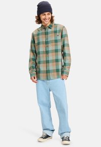 Camisa de franela a cuadros en tonos verdes y marrones, con un cierre de botones en la parte delantera, mangas largas y un bolsillo en el pecho, combinada con jeans azul claro y zapatillas.