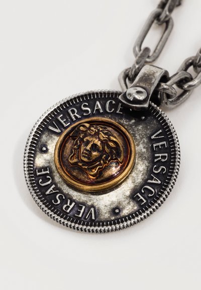 Στρογγυλό μενταγιόν από ασήμι με χρυσή κεφαλή Μέδουσας στο κέντρο, με την εγχάραξη "VERSACE" τρεις φορές γύρω από την άκρη, προσαρτημένο σε μια χοντρή αλυσίδα.