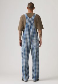 Μπλε και λευκές καρό ολόσωμες φόρμες με τιράντες στους ώμους, μπροστινές τσέπες και χαλαρή εφαρμογή. Κατασκευασμένες από ύφασμα με υφή και μοτίβο.