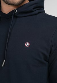 Dunkelblaues Sweatshirt aus Baumwolle mit einem kleinen runden Logo in Rot und Weiß. Kordelzüge mit metallischen Spitzen und einer strukturierten Stoffoberfläche.