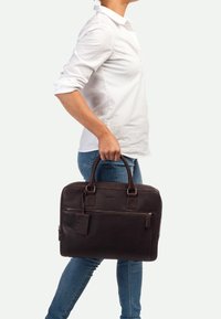 Dunkelbraune Leder-Laptoptasche mit zwei Griffen, Reißverschlussfächern und einem kleinen angebundenen Etikett. Sie hat eine strukturierten Oberfläche und rechteckige Form.