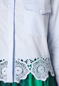 Vaaleansininen napitettavan paidan, jossa on tekstuurinen kangas, paikkatasku ja valkoinen pitsireunus, jossa on pyöreitä kuvioita helman ympärillä.