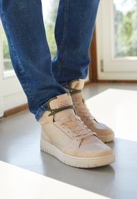 Persoon die beige Tommy Hilfiger hoge sneakers draagt met donkergroene veters en blauwe jeans, staande binnenshuis bij een raam.