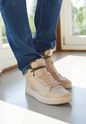 Person wearing beige Tommy Hilfiger high-top sneakers with dark green laces and blue jeans, standing indoors near a window.