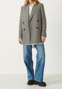 Veston croisé en motif pied-de-poule noir et blanc, avec un col vert et des boutons noirs, porté sur une chemise blanche et un jean bleu.