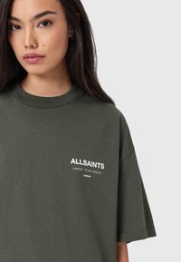 Tummanvihreä ylisuurikokoinen t-paita pehmeästä puuvillakankaasta. Varustettu pyöreällä pääntiellä ja valkoisella tekstipainatuksella "ALLSAINTS" sekä muilla merkeillä edessä.