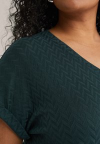 Tyrkysový top s výstřihem do V, který má texturovaný herringbone vzor, krátké rukávy a hladkou, lehkou tkaninu.