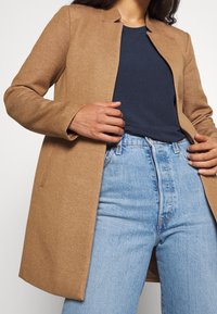 Brun vævet frakke med en kravefri design, lange ærmer og sidelommer, lagdelt over en navy t-shirt og lyseblå højtaljede jeans.