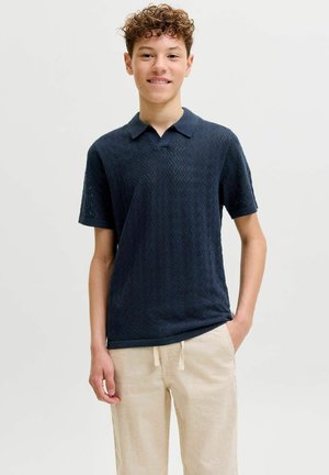 Nooruk, kellel on lokkis juuksed, kandes tumesinist tekstuuriga lühikeste varrukatega polosärki ja beeže nööriga pükse, seisab ühe käega taskus.