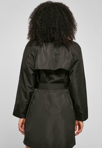 Trench coat verde închis cu talie ajustabilă, mâneci largi și guler lat. Materialul are o textură netedă, ușor lucioasă.