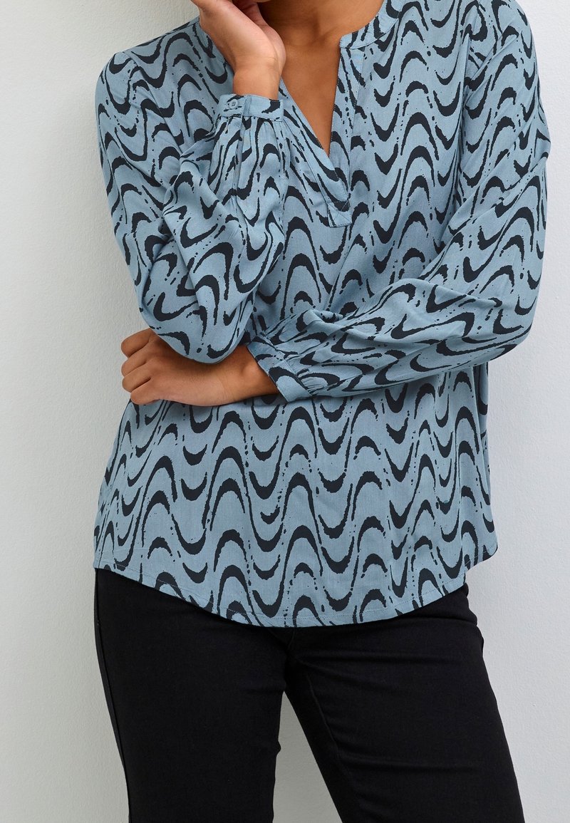 Personne portant une blouse bleu clair à manches longues avec des motifs abstraits noirs en forme de vagues et un pantalon noir, bras croisés et main touchant le menton.