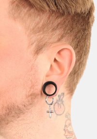 Schwarzer Silikonplug-Ohrstecker mit einem silbernen Kreuzanhänger, der an einem Metallring hängt. Sichtbare Tattoos am Hals und kurzes blondes Haar.