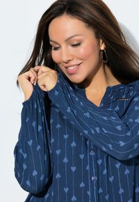 Marineblauwe blouse met een hartjespatroon, knoopsluiting en geplooide mouwen. Gemaakt van zachte stof met een gladde textuur.