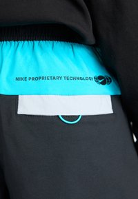 Gros plan sur un pantalon noir avec un rabat de poche blanc, une ceinture élastique turquoise et le texte "NIKE PROPRIETARY TECHNOLOGY" avec logo.