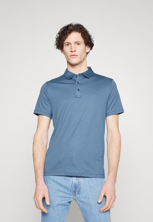 Polo bleu clair en tissu doux, avec un col à boutons, des manches courtes et un logo discret sur la poitrine, associé à un jean clair.