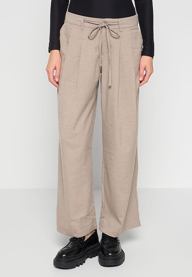 Beige weit geschnittene Hose mit vorderen Falten und Kordelzugbund, kombiniert mit schwarzen schnürbaren Plateauschuhen und einem schwarzen Langarmtop.