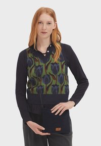 Gilet tricoté avec un fond vert présentant un motif de tulipes bleu foncé ; porté sur un haut à manches longues bleu marine. Sac à bandoulière noir avec un détail de logo.