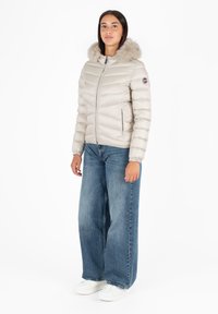 Giacca puffer beige con cappuccio foderato in pelliccia, dotata di una cerniera frontale, polsini elasticizzati e due tasche laterali, abbinata a jeans a gamba larga e sneakers bianche.