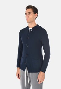Cardigan a maglia blu navy con collo rotondo, chiusura con bottoni e due tasche frontali, caratterizzato da una texture a costine e un design aderente.