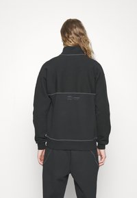 Sudadera negra con cuello alto, puños y dobladillo acanalados, con costuras grises contrastantes y una etiqueta en la parte trasera. Diseño minimalista.