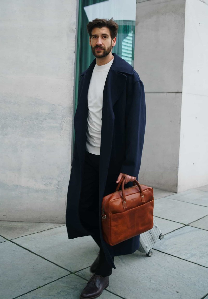 Navy overcoat, white shirt, and dark pants worn by a man holding a brown leather briefcase; gray concrete background with light reflections.