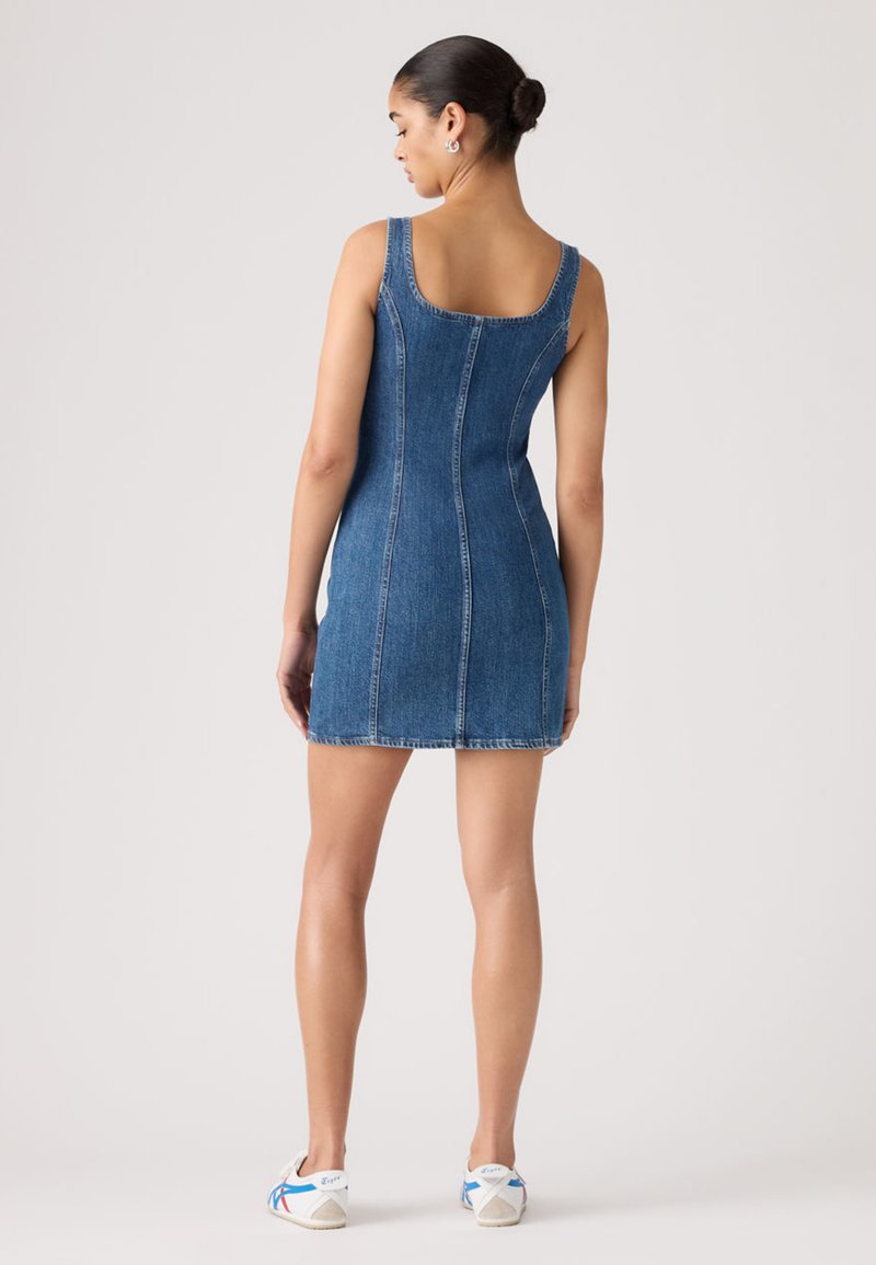 Femme debout, de dos, portant une mini robe en denim bleue sans manches et des baskets blanches avec des détails bleus et rouges.