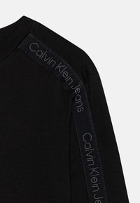 Musta pitkähihainen paita, jossa on teksturoitu kangas ja tummat kaistaleet olkapäillä, joissa on kohokuvioituna "Calvin Klein Jeans" hopeisella.