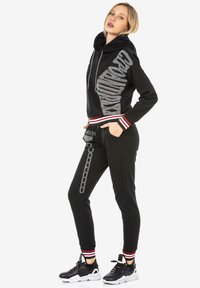 Schwarzer Hoodie mit silbernem Logo, ausgestattet mit Kordelzug und gerippten Bündchen. Kombiniert mit schwarzen Jogginghosen, verziert mit Kettenmuster und roten Akzenten.