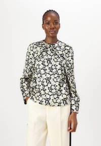 Blumenbluse mit schwarzem Hintergrund, die cremefarbene Blumen, lange Ärmel und einen gerafften Ausschnitt aufweist; kombiniert mit hellfarbigen Hosen.