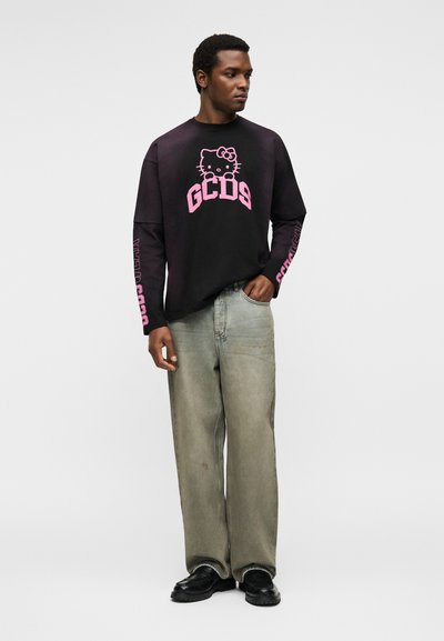 Άνδρας που φοράει μαύρο μακρυμάνικο πουκάμισο με ροζ Hello Kitty και κείμενο GCDS, γκρι φαρδιά τζιν και μαύρα παπούτσια, στέκεται μπροστά σε απλό φόντο.