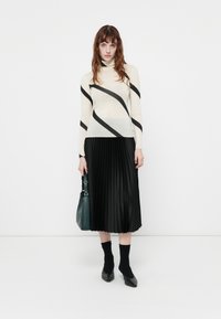 Maglione crema con righe diagonali nere, abbinato a una gonna midi nera plissettata. Gli accessori includono una borsa verde e calzini neri alla caviglia.