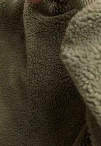 Gros plan d'une personne ajustant le col d'une veste polaire texturée vert brun avec une surface de tissu épaisse et bouclée.