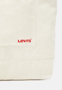 Beżowa torba tote z płótna marki Levi's, z boczną kieszenią i haftowanym na czerwono logo. Wytrzymały materiał z czystymi szwami i wyraźnie zarysowanym kształtem.