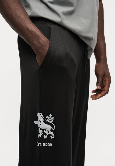 Čierne športové nohavice so bočnými vreckami, s bielou grafikou leva a textom „EST. 2008“. Hladká látka s priliehavým strihom.