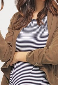 Chaqueta de ante marrón sobre una camisa de rayas azul marino y blanca. Botones visibles en las mangas de la chaqueta. El cabello es ondulado y cae alrededor de los hombros.